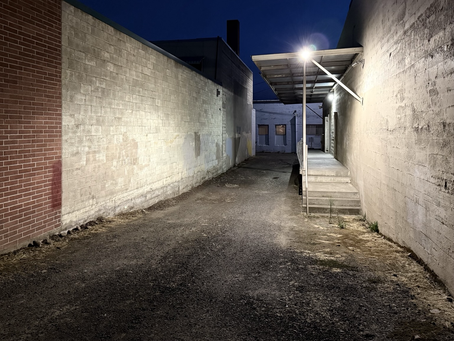 View down empty alley at night, red brick and white painted brick walls.  A glaring bright light over a small loading dock illuminates the alley.
